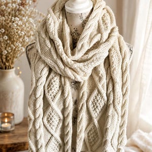 May include: An off-white knitted scarf featuring a complex pattern of vertical cable twists, diamond-shaped textured sections, and seed stitch panels. The scarf is draped around a mannequin and finished with multiple matching yarn tassels at the ends.
