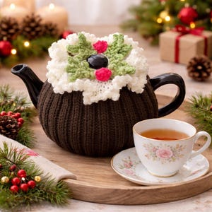 Könnte beinhalten: Eine braune, gestrickte Teekanne mit einem weißen und grünen gehäkelten Stechpalmen-Design. Eine Tasse und Untertasse mit Blumenmuster stehen auf einem Holztablett. Die Teekanne hat einen schwarzen Griff und Ausguss. Festliche Weihnachtsdekoration.