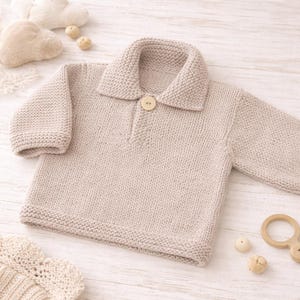 May include: A beige knitted baby sweater with a collared neckline and a single wooden button closure. The long-sleeved sweater has a ribbed hem and cuffs. The image also shows a wooden toy and other baby accessories.