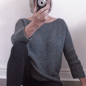 May include: A woman wearing a grey knitted jumper and black leggings. She is sitting on the floor with her legs crossed and holding a camera in front of her face.