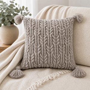 May include: A square, light gray knitted pillow with a cable knit design and four matching tassels. The pillow is placed on a cream-colored blanket and a beige sofa. A vase with greenery is in the background.
