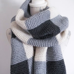 Może przedstawiać: Wełniany szalik w kratkę w odcieniach szarości i bieli. Szalik jest złożony i drapowany na białym manekinie.