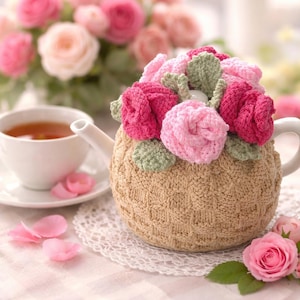 Könnte beinhalten: Eine beige Teekanne mit einer gestrickten Hülle, die mit gehäkelten rosa und fuchsiafarbenen Rosen und grünen Blättern verziert ist. Eine weiße Tasse und Untertasse mit Tee und Rosenblättern befinden sich in der Nähe, mit einem Rosenstrauß im Hintergrund.