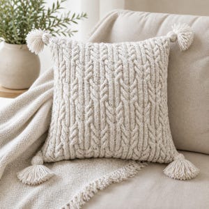 Puede incluir: Cojín tejido de color crema con un diseño de punto de cable y cuatro borlas decorativas. El cojín cuadrado descansa sobre un sofá beige con una manta a juego. Un jarrón con vegetación está en el fondo.
