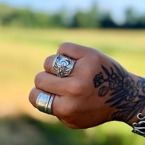 May include: A silver ring featuring a crescent moon and star motif with intricate embossed scrollwork details, worn on the ring finger alongside a wider, plain silver band on the middle finger, both displaying a polished metallic finish.