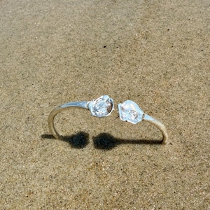 May include: A silver-colored open cuff bracelet with two decorative ends, each featuring a clear, faceted stone. The bracelet is resting on a sandy surface, with its shadow visible below. The design is simple and elegant.