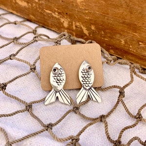 May include: A pair of silver-toned fish-shaped stud earrings featuring engraved scale patterns and a small dark circular accent for the eye. The earrings are attached to a rectangular brown card, displaying a detailed metallic finish and pointed tail fins.