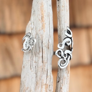 May include: Two silver-colored rings with ornate, swirling designs. One ring features a floral-like design, while the other has an open, abstract pattern. The rings are displayed on a weathered wooden surface, showcasing their intricate details.