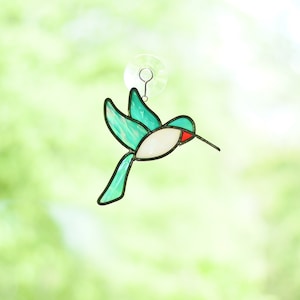 Op de afbeelding: Een gebrandschilderde kolibrie met turquoise vleugels en staart, een wit lichaam en een rode snavel. De vogel is zwart omlijnd en wordt opgehangen door een transparante zuignap. De achtergrond is een zacht, wazig groen.