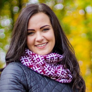 Puede incluir: Una mujer con el pelo largo y oscuro lleva una bufanda infinita de color morado y blanco con un patrón. Está sonriendo y mirando a la cámara. El fondo está desenfocado.