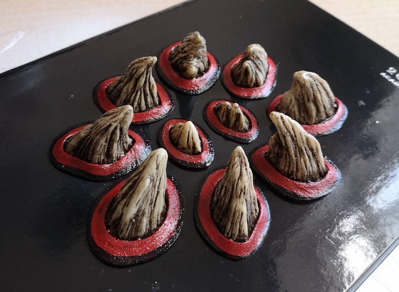 May include: Nine small, pointed, beige-colored horns with a rough texture are arranged in a circle on a black surface. Each horn is surrounded by a red ring.