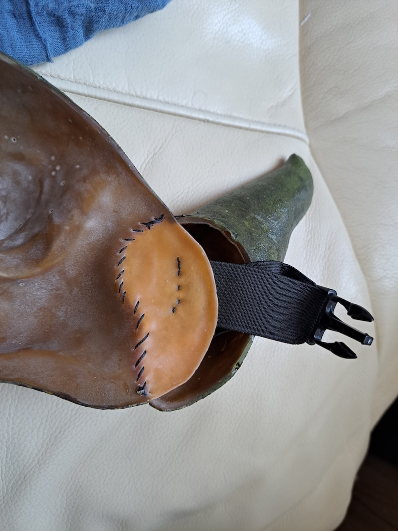 May include: A brown and green mask with a black elastic strap and buckle. The mask has a stitched leather-like appearance with a rounded shape. The strap is attached to the mask with a buckle for adjustment.