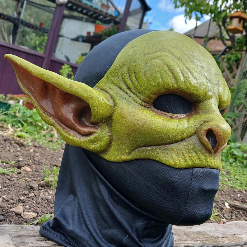 May include: A green goblin mask with large ears and a textured surface. The mask has a black hood and is designed to cover the face. The mask is likely made of a durable material and is suitable for costume or cosplay.