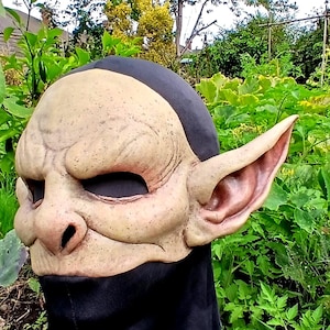 Peut inclure: Un masque recouvrant toute la tête présentant un visage beige texturé rappelant un gobelin, avec des oreilles pointues saillantes, des orbites profondes et un nez large. Le masque est fixé à une cagoule en tissu noir couvrant le cou.