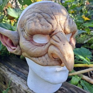 May include: A detailed goblin mask with a textured, weathered appearance. The mask is primarily tan with hints of red and white, featuring prominent wrinkles, a long nose, and pointed ears. It is displayed on a white foam head.