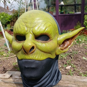 May include: A green goblin mask with large ears, a prominent nose, and black eye openings. The mask is worn over a black fabric covering the neck and lower face. The mask has a textured, weathered appearance.