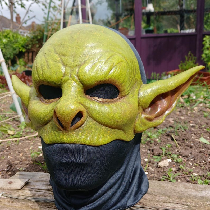 May include: A green goblin mask with large ears, a prominent nose, and black eye openings. The mask is worn over a black fabric covering the neck and lower face. The mask has a textured, weathered appearance.