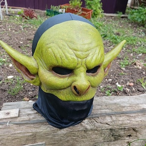 May include: A green goblin mask with large pointed ears, a wrinkled face, and black eye holes. The mask is made of a textured material and is attached to a black hood. The mask is resting on a wooden surface.