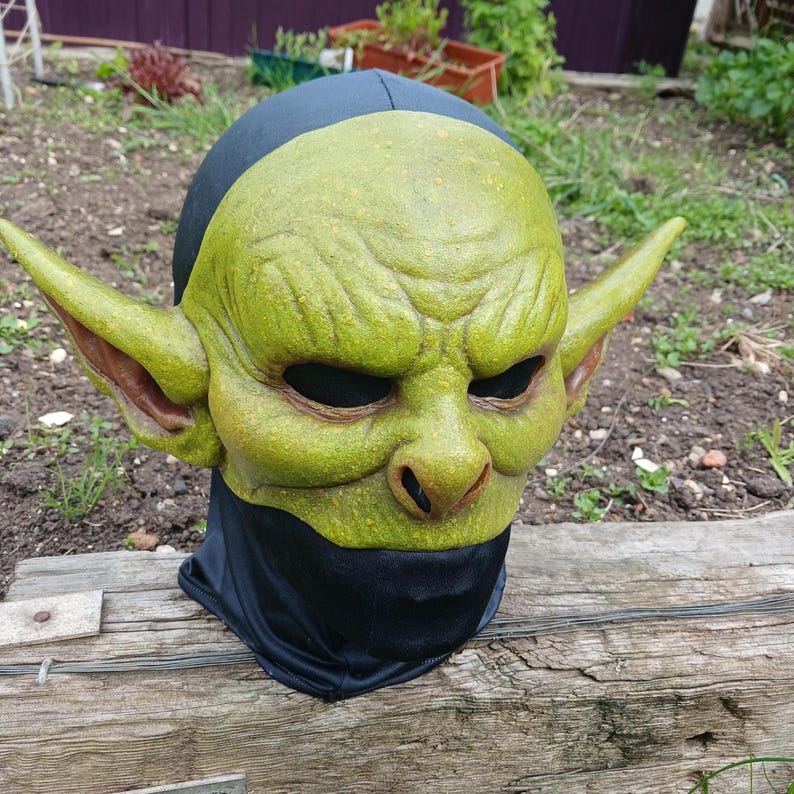 May include: A green goblin mask with large pointed ears, a wrinkled face, and black eye holes. The mask is made of a textured material and is attached to a black hood. The mask is resting on a wooden surface.