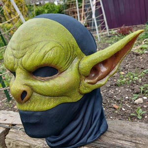 May include: A green goblin mask with large ears and a textured surface. The mask has dark eye holes and is worn over a black fabric covering the lower face and head.