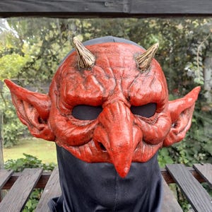 Op de afbeelding: Een rood duivelsmasker met twee puntige hoorns op het voorhoofd, grote puntige oren aan de zijkanten, een prominente haakneus en diepliggende oogopeningen. Het oppervlak heeft een getextureerd, verweerd uiterlijk met donkere schaduwen.