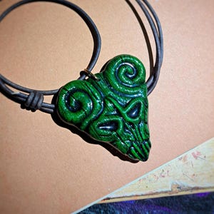 May include: A green pendant necklace with a heart-shaped design. The pendant has a detailed, textured pattern with dark green and black accents. The necklace has a black cord.