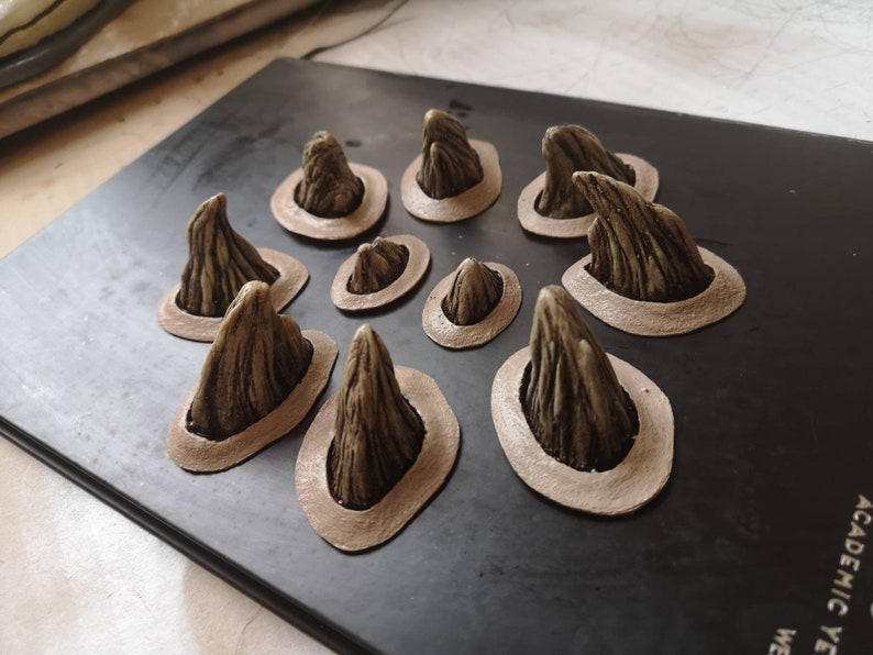 May include: Ten small, detailed, beige, resin, sculpted, pointy, horn-like shapes, each with a textured surface, are arranged in a circle on a black surface. Each horn is set in a circular, brown, raised, decorative frame.