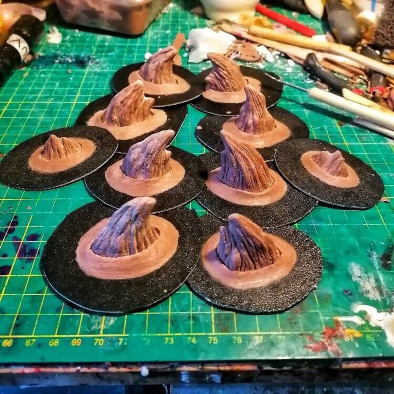 May include: A collection of ten brown witch hat toppers with a pointed, textured design. The toppers are arranged on a green cutting mat with black markings.