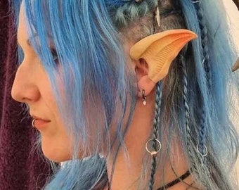 May include: A person with blue hair wearing a pair of elf ears. The ears are made of a light brown material and are attached to the person's ears with a small metal ring.