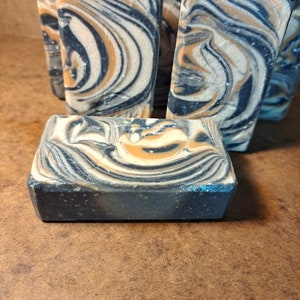 Op de afbeelding: Een stapel handgemaakte zeepstaven met een swirlend zwart en oranje patroon. De zeepstaven zijn rechthoekig en hebben een ruwe textuur.