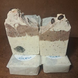 May include: Three bars of handmade soap wrapped in white paper. The soap is brown, white, and beige with coffee beans embedded in the soap. The soap is labeled "6.0 oz (170 g)".