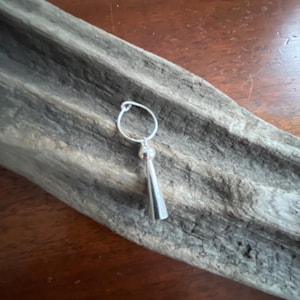 May include: A silver-toned earring featuring a thin circular hoop attached to a small spherical bead and a smooth, elongated conical pendant. The metal has a polished, reflective finish, creating a clean and simple design for this jewelry piece.