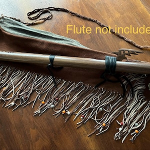 May include: A wooden flute with a leather strap and a decorative bird carving. The flute rests on a brown leather case with fringe and colourful beads. The text "Flute not included" is visible.