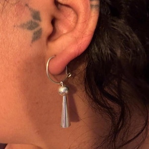 May include: Silver hoop earrings with a dangling cone-shaped charm. The earrings are worn in the left ear.