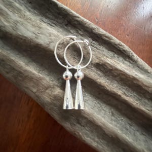 May include: A pair of silver hoop earrings featuring a dangling charm composed of a small polished sphere attached to a slender, elongated cone shape. The earrings have a bright, reflective metallic finish and a simple, clean design structure.