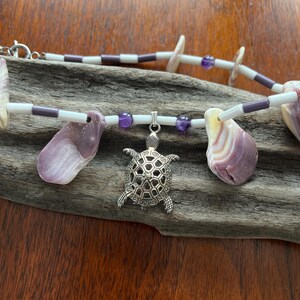 Könnte beinhalten: Eine Halskette aus weißen und violetten Perlen, Muscheln und einem silbernen Schildkrötenanhänger. Die Halskette wird auf einem Stück Treibholz präsentiert.