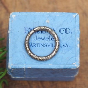 May include: A silver-toned ring with an engraved floral pattern rests on a light blue box. The box has text that reads "BYRD JEWELERS & CO. MARTINSVILLE, VA." A sprig of rosemary is in the lower left corner.