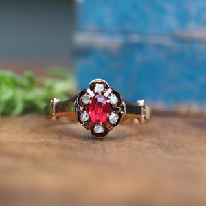 May include: A gold ring featuring a central oval-cut red gemstone surrounded by a cluster of small, clear, round-cut stones set in a dark metal frame. The band is made of polished gold and has a smooth, tapered design leading to the setting.
