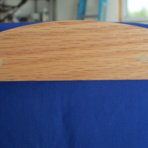 May include: A close-up of a wooden object with a curved top edge, featuring a light brown color and visible wood grain. Two small, light-colored circular pegs are visible. The object is set against a backdrop of deep blue fabric.
