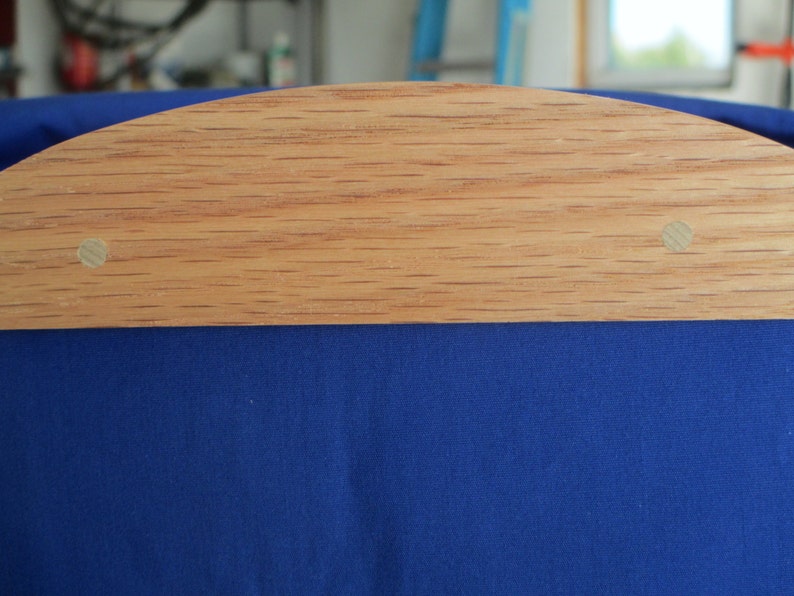 May include: A close-up of a wooden object with a curved top edge, featuring a light brown color and visible wood grain. Two small, light-colored circular pegs are visible. The object is set against a backdrop of deep blue fabric.