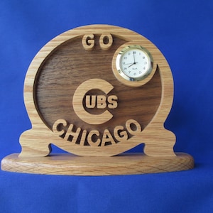 May include: Wooden desk clock with the words "GO CUBS CHICAGO" carved into the wood. The clock face is round with a gold-colored bezel. The clock is made of light and dark brown wood.