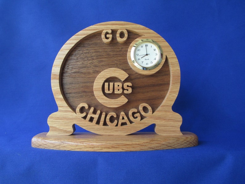 May include: Wooden desk clock with the words "GO CUBS CHICAGO" carved into the wood. The clock face is round with a gold-colored bezel. The clock is made of light and dark brown wood.
