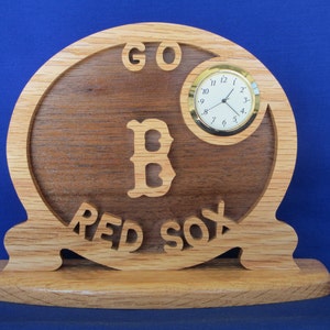 May include: Wooden clock with the words "GO RED SOX" and a "B" logo carved into the wood. A small clock face with gold trim is set into the wooden design. The clock is made of light and dark brown wood.