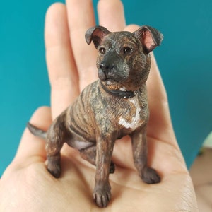 Puede incluir: Una pequeña y detallada figura de un perro con pelaje atigrado marrón y negro. El perro está sentado con las orejas levantadas y lleva un collar negro. La figura se sostiene en una mano.