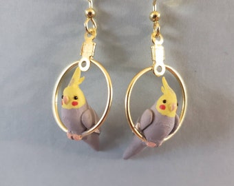 May include: A pair of gold-colored hoop earrings, each featuring a miniature, hand-painted cockatiel. The birds are gray with yellow crests and red cheek patches, perched inside the hoops. The earrings have a simple hook closure.