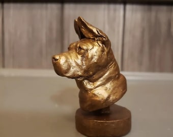 Puede incluir: Un busto de cerámica de color dorado de la cabeza de un perro. El perro es de una raza con orejas grandes y caídas y una cara arrugada. El busto está sobre una base redonda.