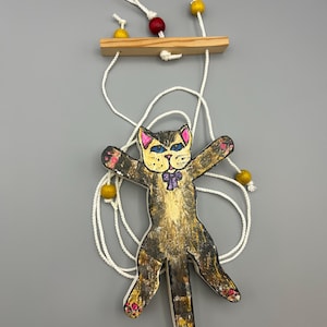 May include: A whimsical cat-shaped wooden ornament with outstretched arms, painted in shades of brown, black, and gold. The cat has a purple bow tie and pink accents on its paws. It hangs from a wooden bar with white string and yellow and red beads.