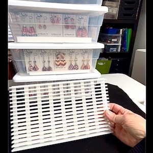 Peut inclure: Plusieurs boîtes de rangement en plastique transparent empilées, remplies de présentoirs de boucles d'oreilles. Les boucles d'oreilles sont sur des cartes blanches. Une grille blanche est tenue au premier plan.