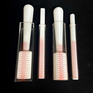 Può includere: Quattro strumenti medici trasparenti in plastica e bianchi con segni rossi. Due strumenti hanno una parte superiore bianca bulbosa e sono racchiusi in un tubo di plastica trasparente. Gli altri due sono rettangolari con linee rosse.