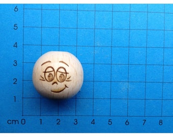 May include: A wooden bead with a hand-drawn face. The face has closed eyes, a small smile, and eyelashes.